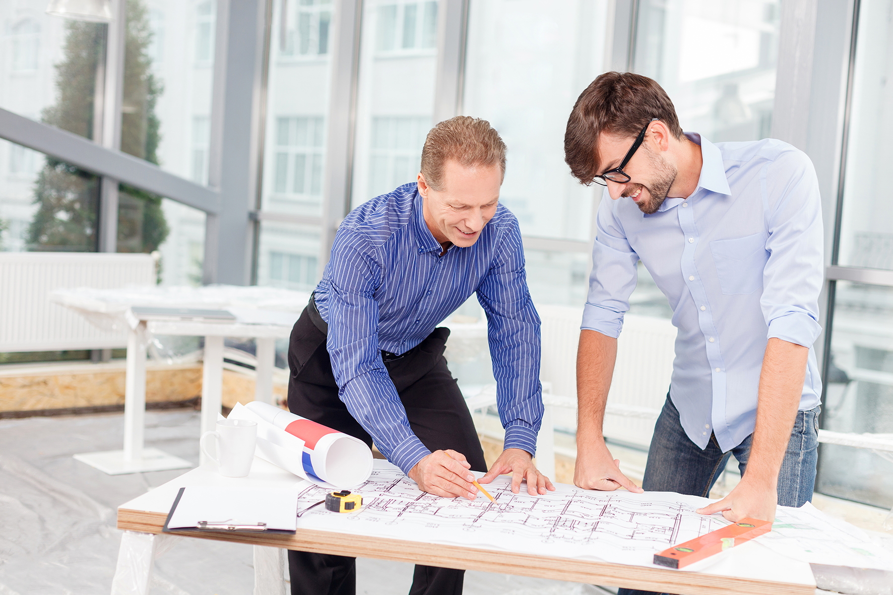 Cheerful senior architect is explaining to his young colleague the plan of building. The men are looking at the blueprint on the table with aspiration. They are standing and smiling